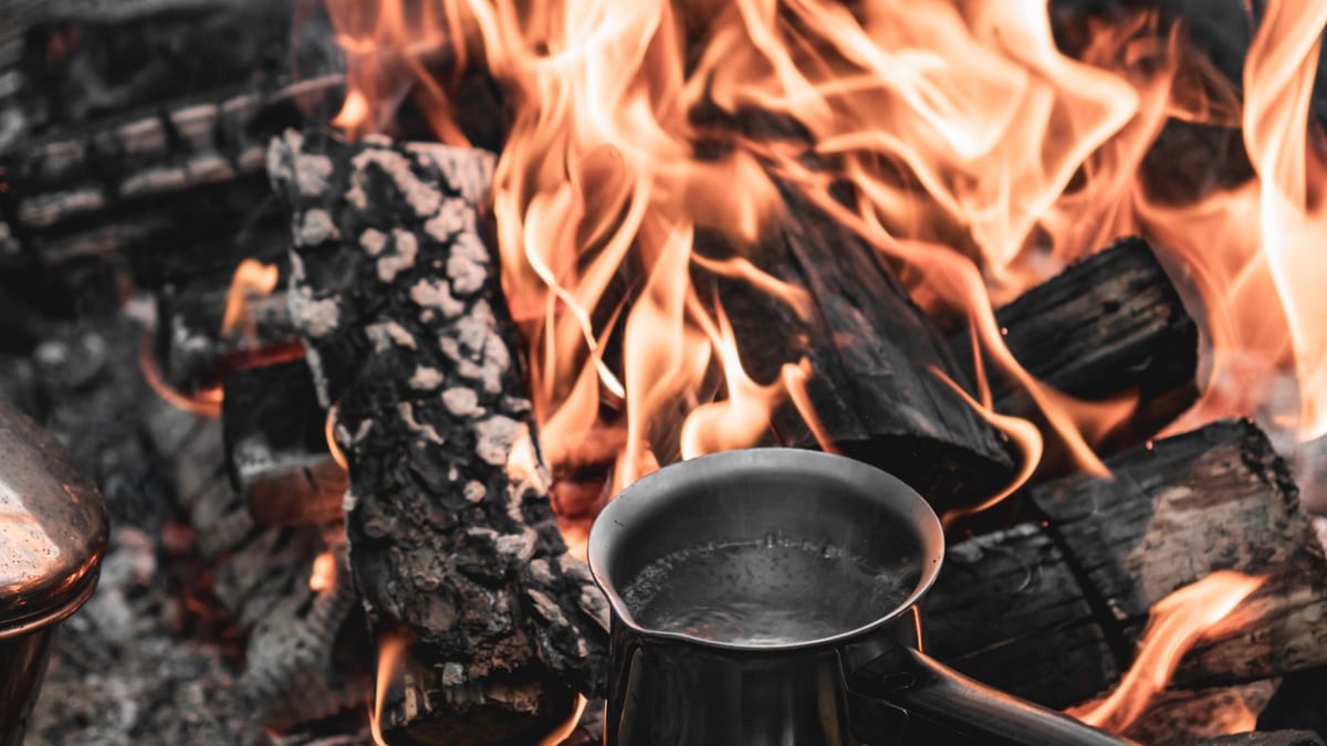 Metal coffee pot heating beside open campfire flames and charred logs